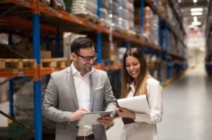 Two colleagues review a tablet and paperwork beside warehouse shelves.