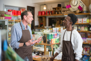 Two colleagues in aprons talk together in a neighborhood grocery store.