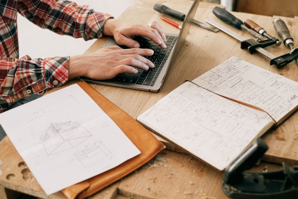 Hands using a laptop beside project plans and tools on a workshop table